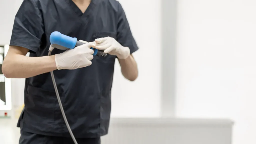 A practitioner wearing scrubs and gloves adjusts a shockwave therapy handpiece during a treatment session.